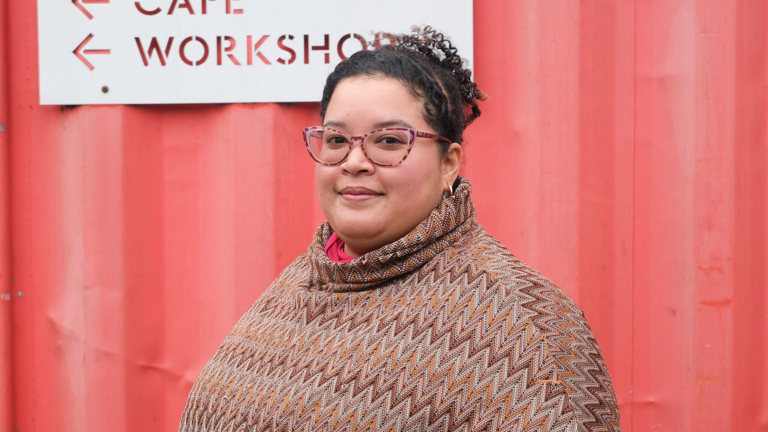 Female reception and office manager against red container wearing patterned top and glasses