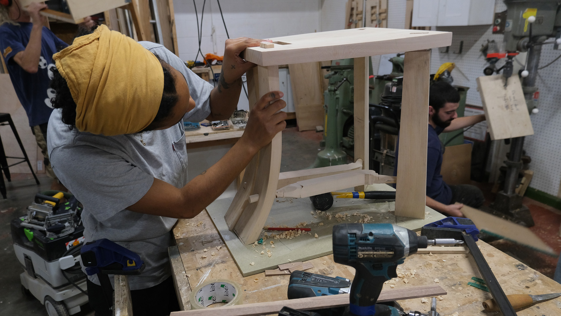 Woman with yellow headscarf building a chair on a woodwork bench