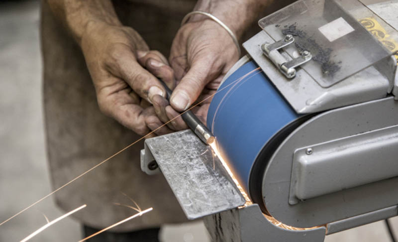 two hands guiding a metal rod over a metal belt sander