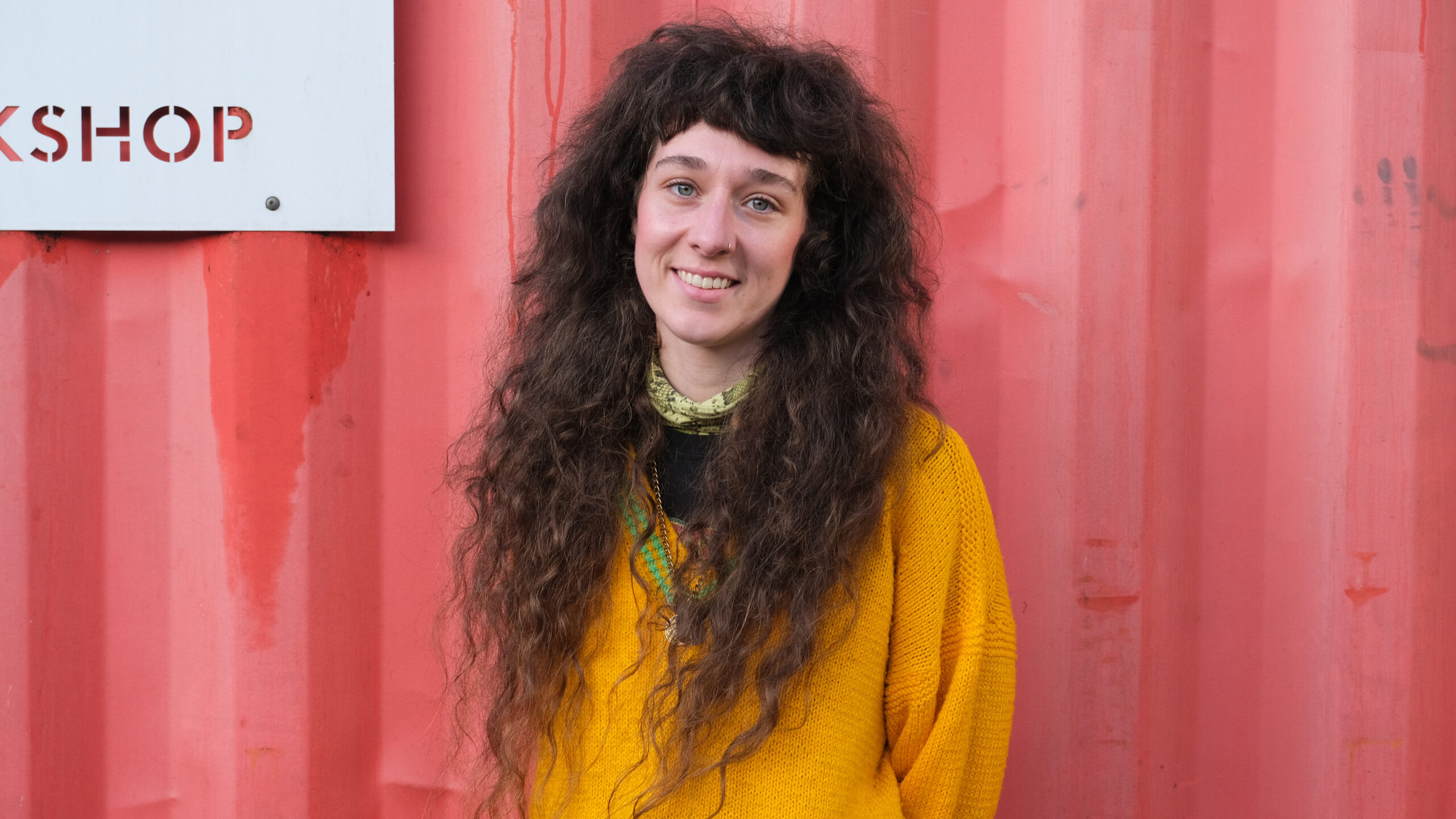 staff member with long brown hair stood against red container wearing yellow knitted jumper
