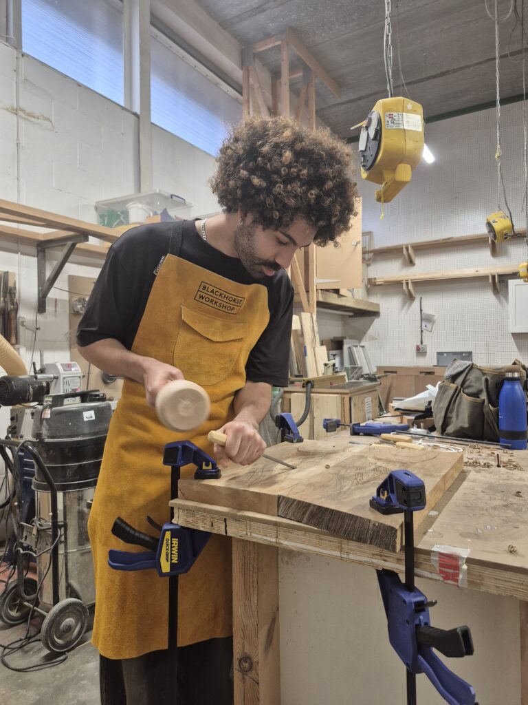 young man in a workshop carving in to wood wearing a yellow Blackhorse workshop branded apron