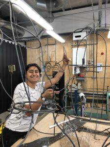 fawzy in the picture with short hair, in a workshop setting holding a sculture that is they size of a sofa make from metal bent bars